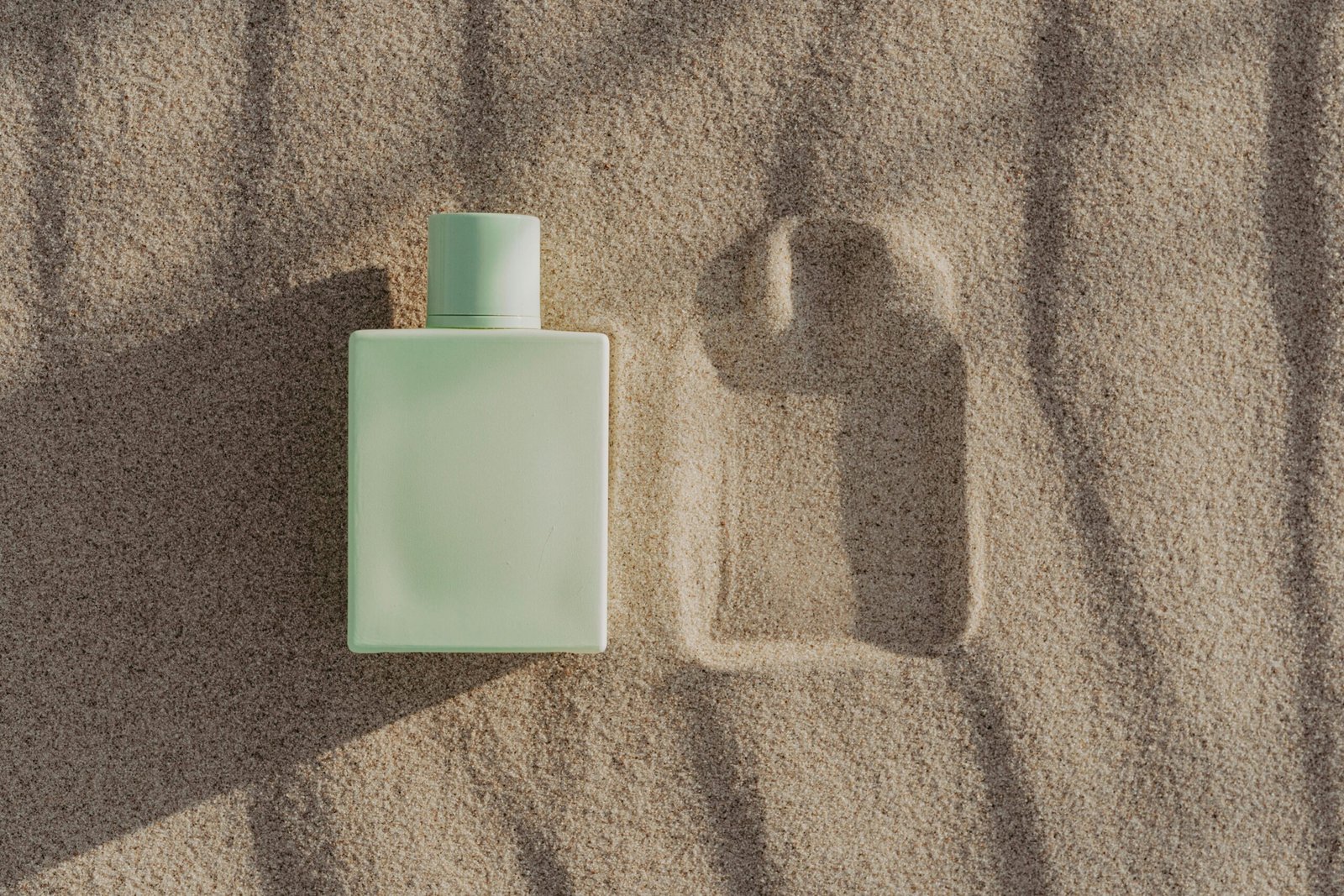 O sticlă dreptunghiulară de parfum verde deschis pe nisip, care prezintă umbra sa și o amprentă - un joc artistic de creație, perfect pentru imagini de marketing surprinzătoare.